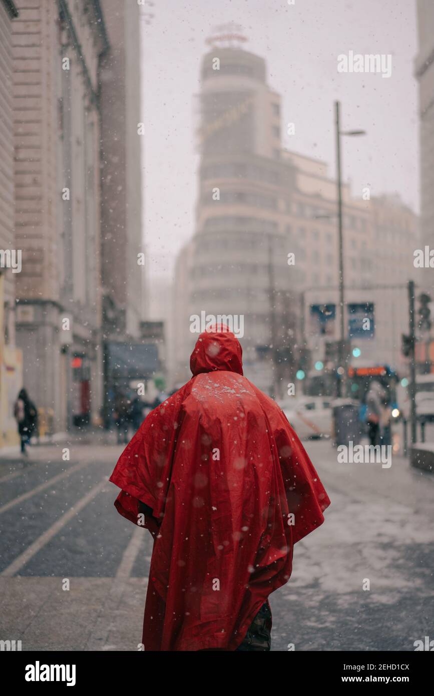 Rückansicht des anonymen Mannes in leuchtend rotem Schutzmantel Spaziergang auf der Straße mit Gebäuden im verschneiten Winter in Madrid Spanien Stockfoto