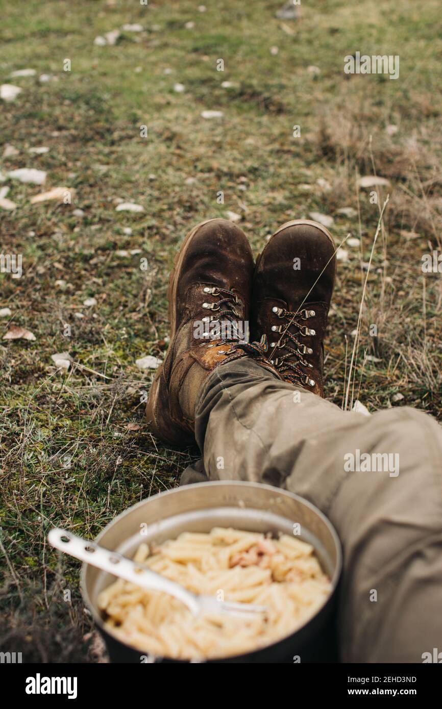 Crop anonymen männlichen Reisenden in Stiefeln sitzen auf dem Boden mit Schale Makkaroni während der Reise in die Natur Stockfoto