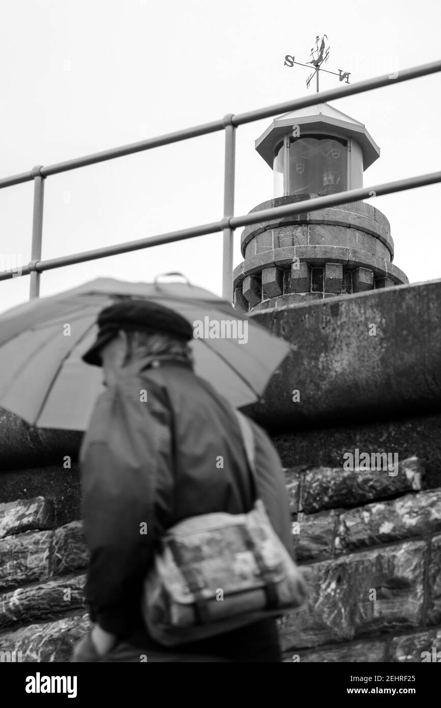 Mann mit Schirm, der an Windfahne ( Anemometer ) im Winter in Schwarzweiß vorbeiläuft, Teignmouth, Devon, England Stockfoto