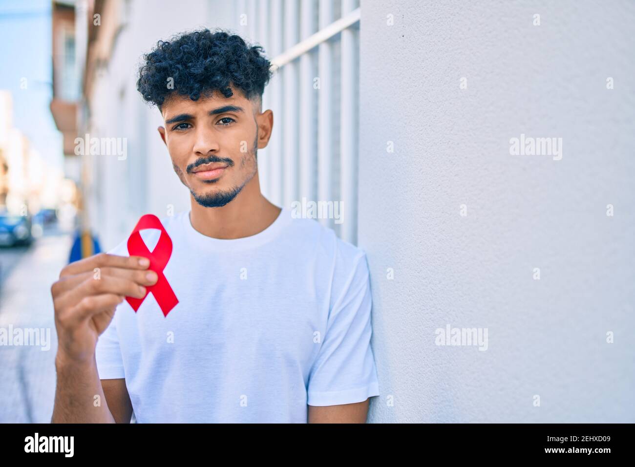 Junger arabischer Mann mit ernstem Ausdruck, der an der Wand gelehnt ein rotes Band mit hiv-Bewusstsein hält. Stockfoto
