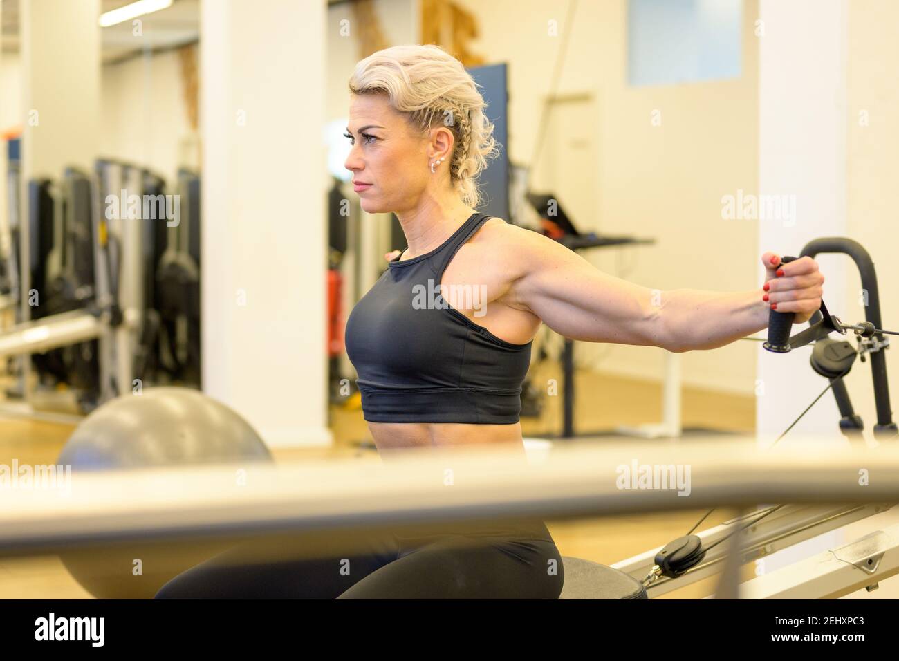 Sportliche Frau mittleren Alters, die an der Widerstandsausrüstung in einem arbeitet Gym Stärkung ihrer Arm und Oberkörper Kernmuskeln angesehen An anderen Geräten in vorbei Stockfoto