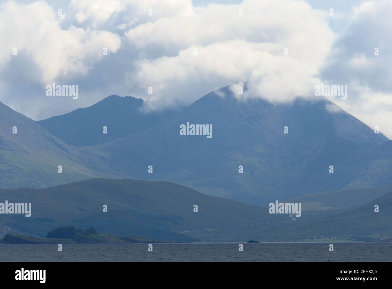 Weit entfernte Straße über dem Wasser windet sich zu einem riesigen felsigen Berg auf einer schottischen Insel. Helle, sonnenbeschienenen Wolken bedecken den Gipfel des Berges. Querformat. Stockfoto