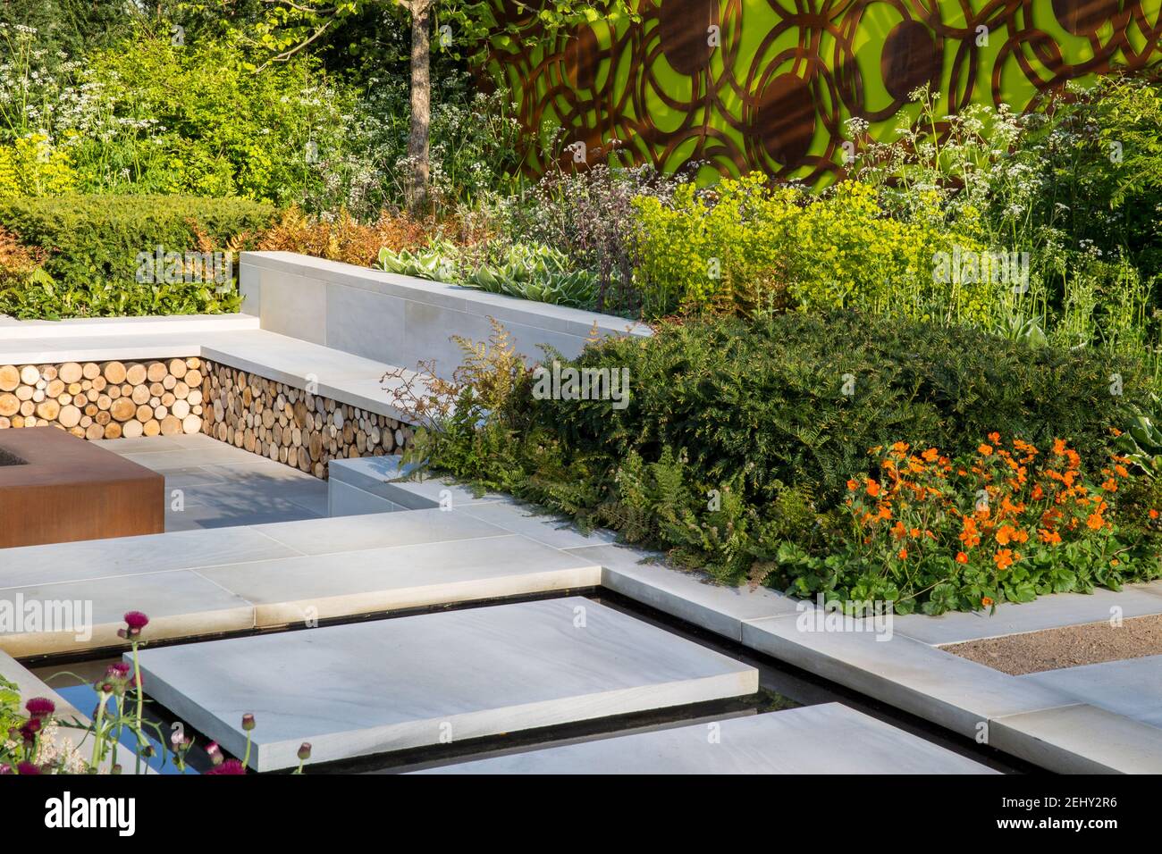 Blick auf den modernen englischen Garten aus Yorkstone mit Steinplatten über einem Wasserspiel, der zu einem tiefer gelegenen Garten mit Steinbänken führt, die an das Frühjahr Großbritannien Grenzen Stockfoto