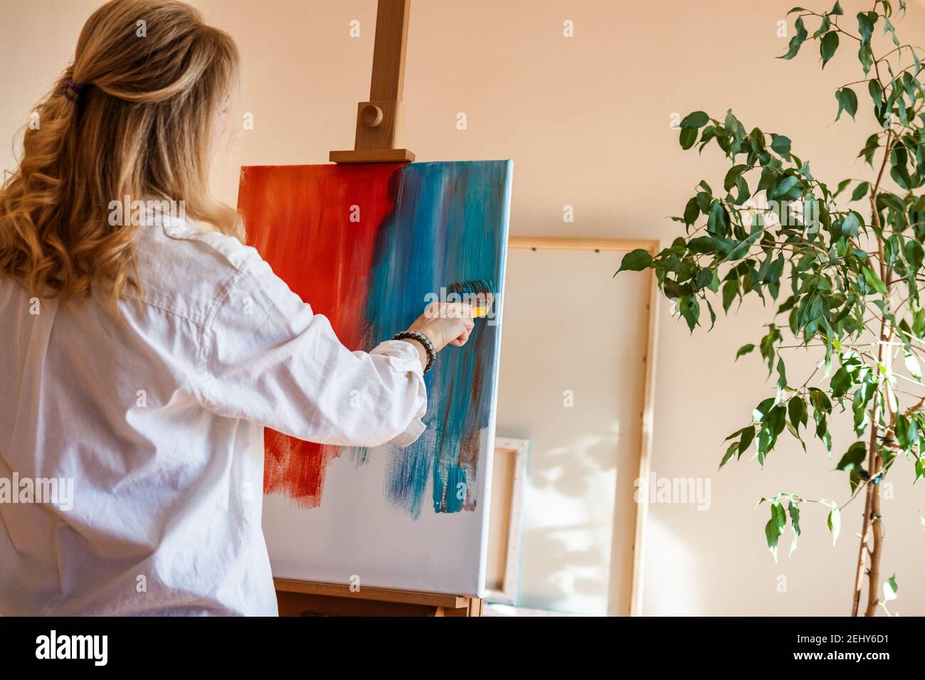 Frau arbeitet an zeitgenössischen abstrakten Ölgemälde im kreativen Studio. Blonde Haare kaukasischen Maler stehen mit Pinsel und Staffelei drinnen Stockfoto