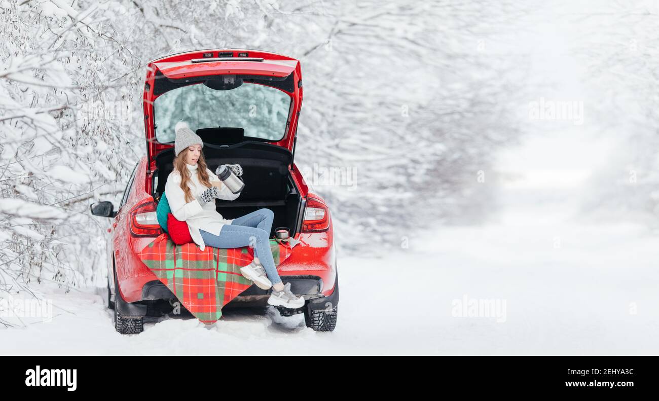 Die junge Frau gießt ein heißes Getränk, Kaffee, Kakao oder Tee aus einer Thermoskanne, während sie im Kofferraum eines roten Autos auf dem Hintergrund eines Winterwaldes sitzt Stockfoto