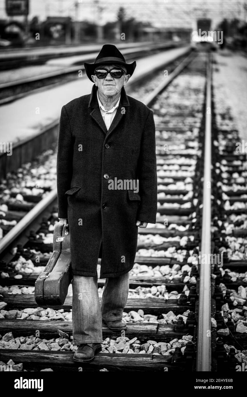 Ein Mann in einem schwarzen Mantel mit einem schwarzen Hut Und schwarze Sonnenbrille und eine Gitarre in der Hand beim Gehen Die Bahnstrecke hinunter Stockfoto