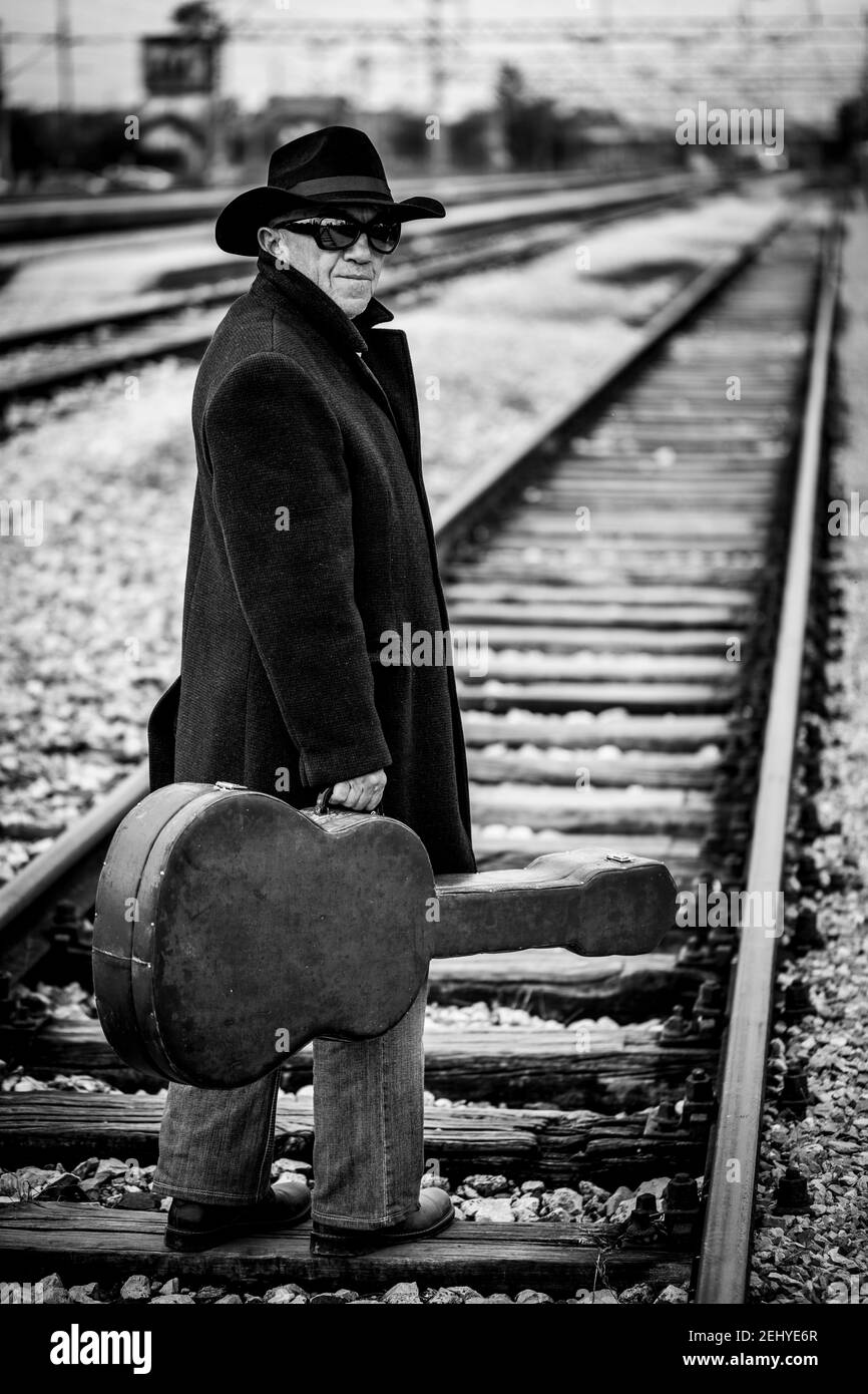 Ein Mann in einem schwarzen Mantel mit einem schwarzen Hut Und schwarze Sonnenbrille und eine Gitarre in der Hand beim Gehen Die Bahnstrecke hinunter Stockfoto