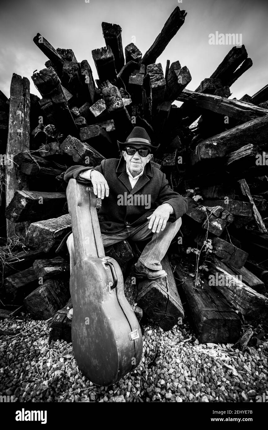 Ein Mann in einem schwarzen Mantel mit einem schwarzen Hut Und schwarze Sonnenbrillen sitzen neben schwarzen Holzbalken mit Seine Gitarre Stockfoto