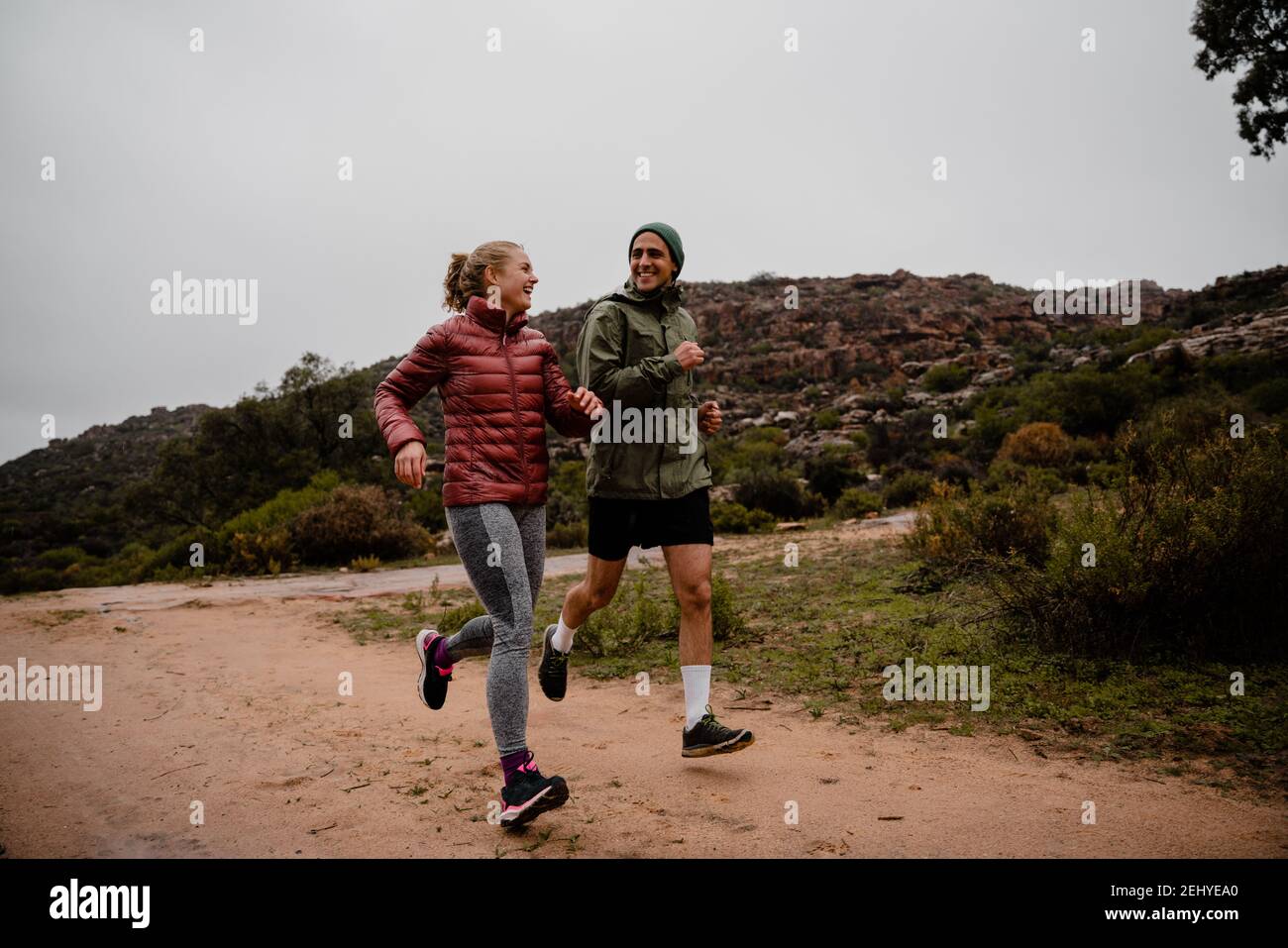 Junges fittes Paar lachend beim Laufen auf Schotterbergbahn Bei bewölktem Wetter mit üppigen Büschen Stockfoto