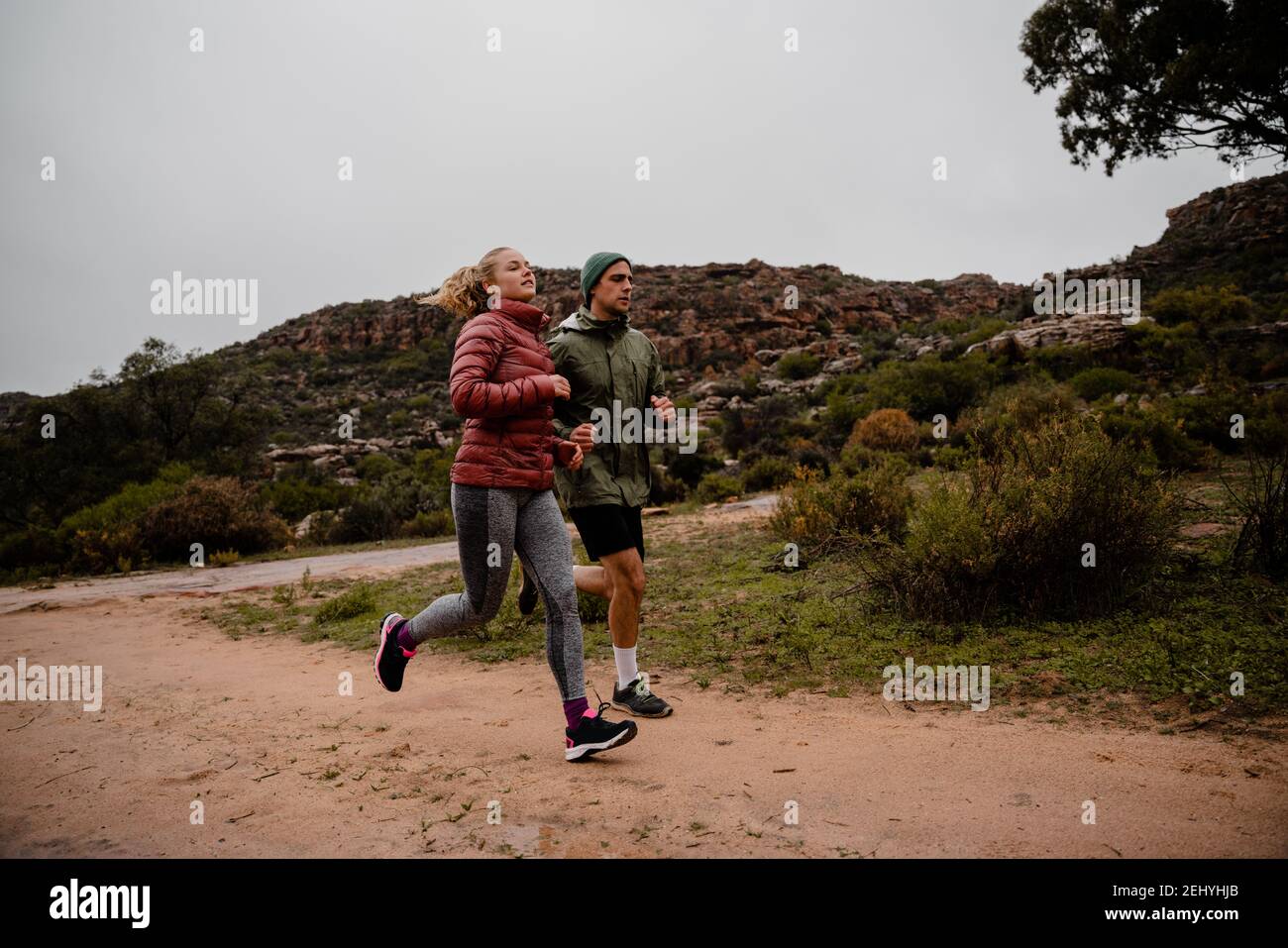 Starke Passform junge männliche und weibliche Athleten Trail Running on Kalte Winter Tag auf Bergweg bewölktes Wetter und Büsche Stockfoto
