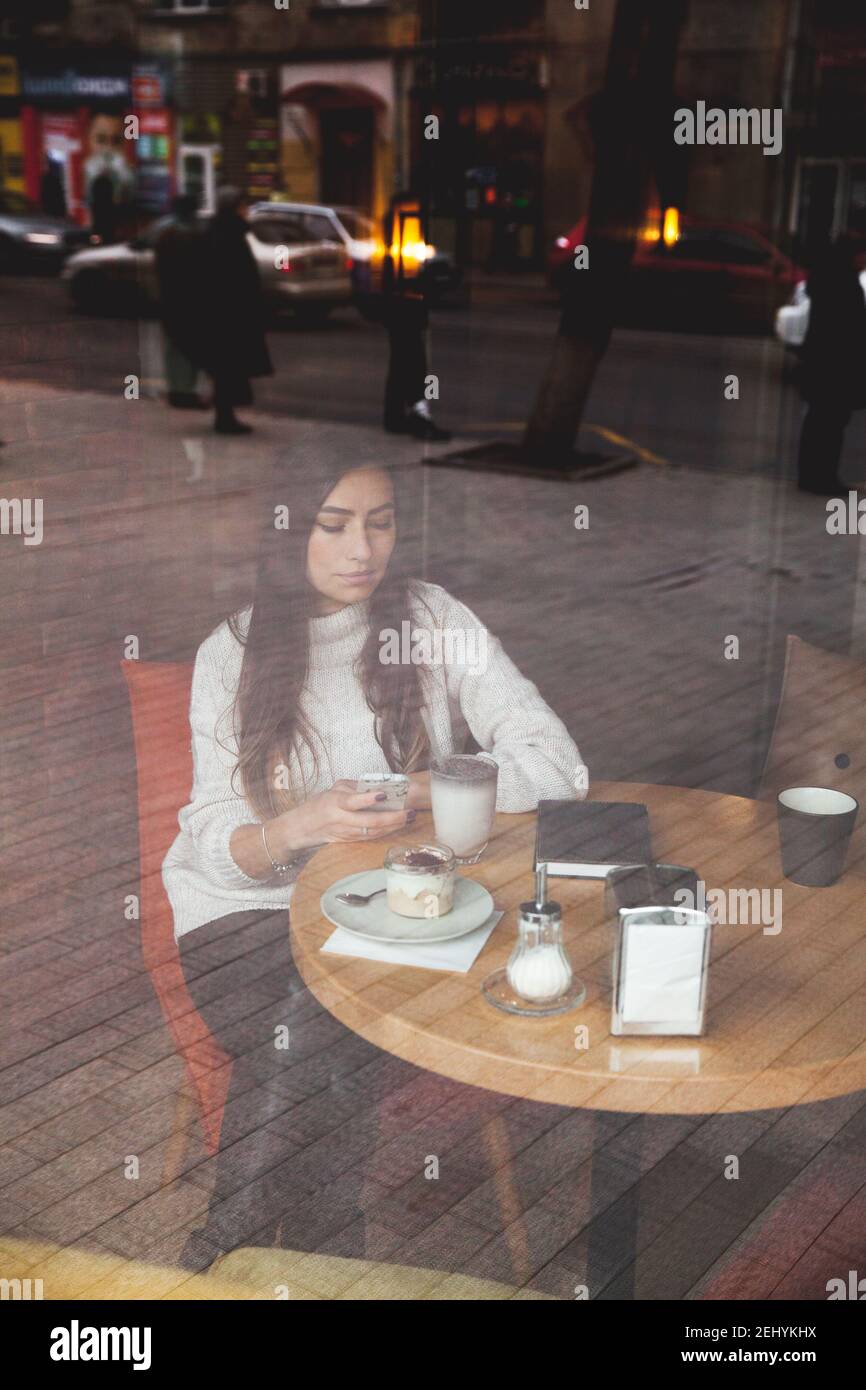 Charmante Frau schaut am Telefon im Café. Leckere Schokoladenkuchen und Kaffee auf dem Tisch. Heller sonniger Morgen im Café A Stockfoto