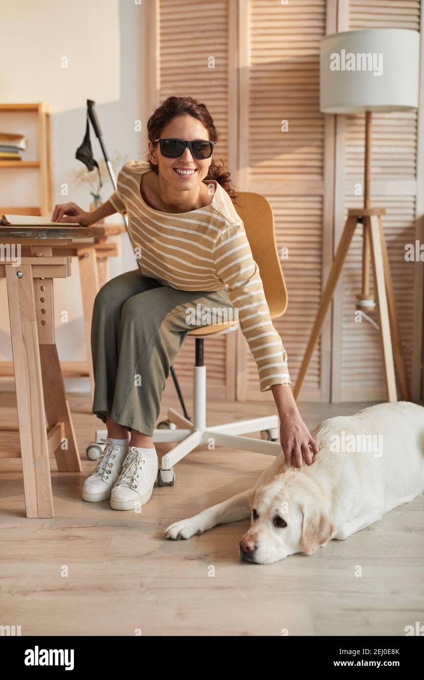 Vertikale voller Länge Porträt der modernen blinden Frau sitzen an Tisch im gemütlichen Haus Interieur und Streichelführer Hund Stockfoto