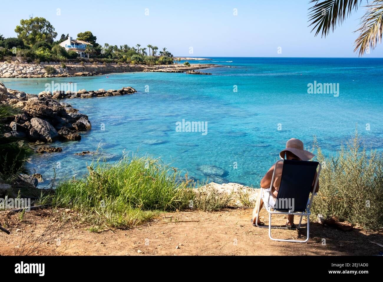 Frau sitzt auf einem Stuhl und entspannt ein Buch lesen während der Sommerferien am Strand mit türkisfarbenem ruhigem Wasser. Stockfoto