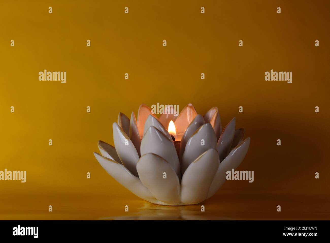 Brennende Teelicht mit Flamme in einem keramischen Lotusblumen Kerzenhalter auf einem tiefgelben Hintergrund mit dramatischer, romantischer Beleuchtung und Kopierraum Stockfoto