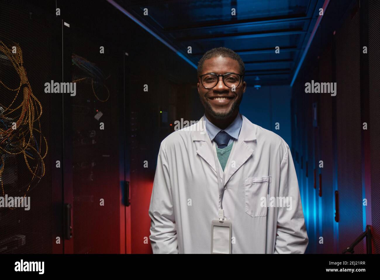 Porträt eines afroamerikanischen Data Scientists in Labormantel und lächelnd an der Kamera, während er mit Supercomputer im Serverraum arbeitet, Kopierraum Stockfoto