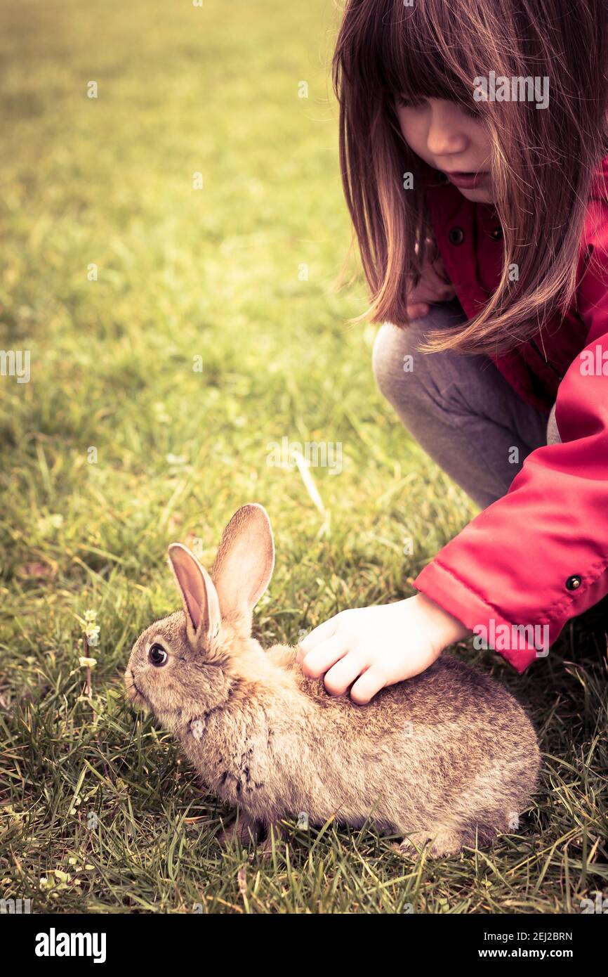 Kleines Kind Mädchen, trägt rote Jacke, Kuscheln Osterhase im Gras. Frühlingshintergrund, Hintergrundbild mit Kopierraum. Vintage-Töne. Vertikale Aufnahme. Stockfoto