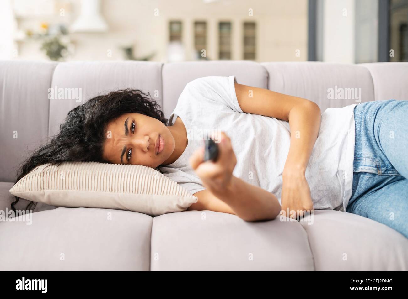 Aufgeregt junge Birazialfrau liegt auf einem weißen Sofa mit einer Fernbedienung in der Hand, kann nichts Interessantes im fernsehen finden, auf einer Selbstisolation, zu Hause Stockfoto