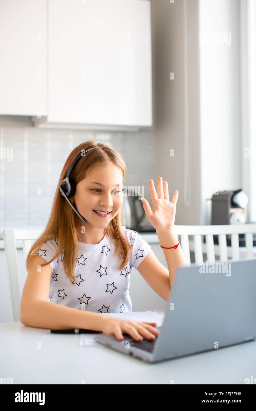 Ein Mädchen im Headset vor einem Laptop-Monitor winkt vor einer Online-Lektion Hand zu einem Lehrer. Online-Bildungskonzept. Vertikales Foto. Stockfoto