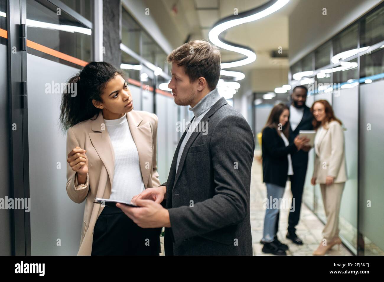Zwei multirassische erfolgreiche Kollegen kommunizieren in einem modernen Büro, diskutieren neue Ideen, denken über einen Plan nach, im Hintergrund ihre Kollegen Stockfoto
