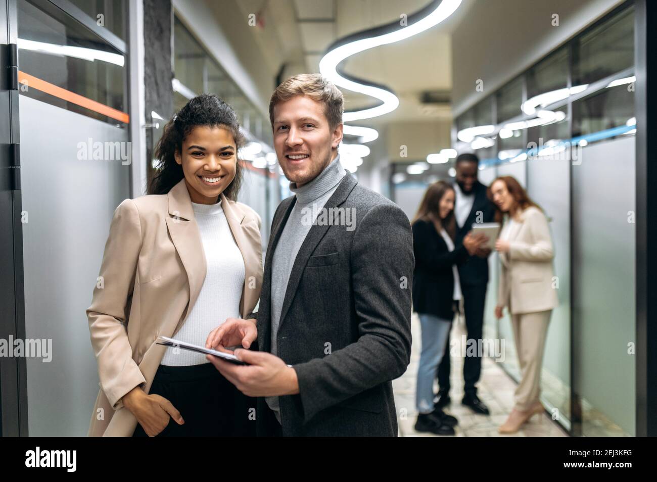 Porträt von zwei multirassischen zufriedenen erfolgreichen Kollegen in formeller Kleidung, die in einem modernen Büro stehen und freundlich lächeln auf die Kamera Stockfoto