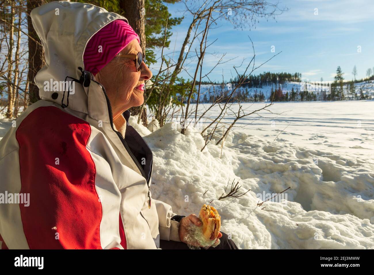 Ältere Frau, die sich ausruhte, sitzt am Ufer eines gefrorenen Sees und hält ein Sandwich in der Hand, Bäume und einen blauen Himmel im Hintergrund, Bild von Vastre Stockfoto