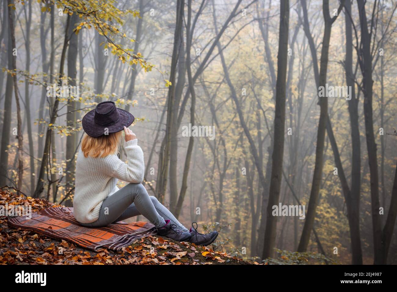 Frau entspannen in Nebelwald in der Herbstsaison. Frau trägt Strickpullover und Hut sitzt auf Decke im Freien. Genuss der Natur Stockfoto