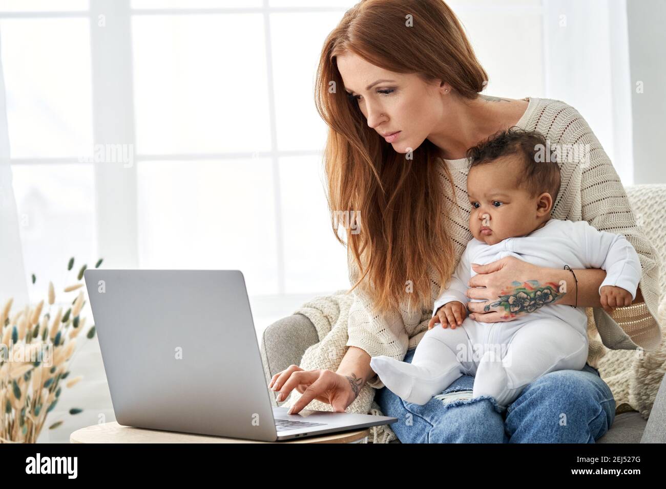 Vielbeschäftigte Mutter mit Laptop, die von zu Hause aus mit kleinen afrikanischen Baby-Tochter arbeitet. Stockfoto