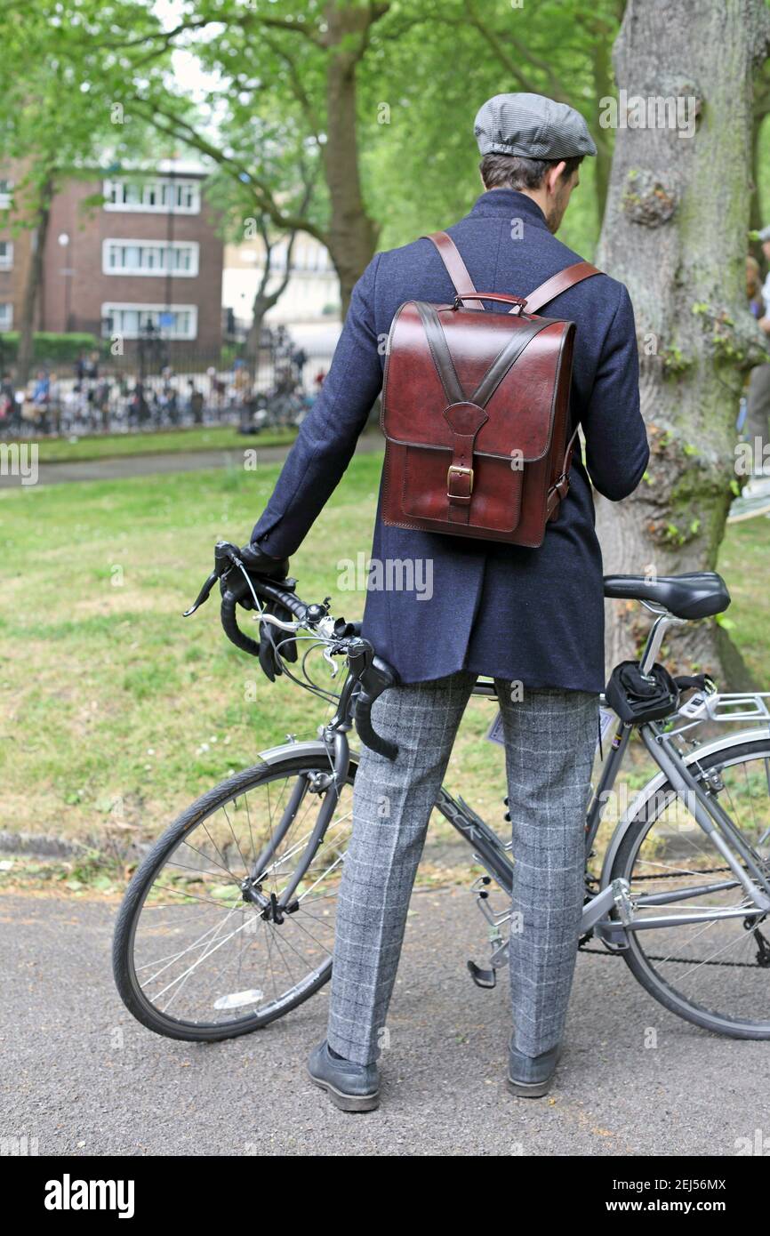 Ein hübscher Kaukasusmann, der sein Fahrrad in Park hält und in London, Großbritannien, elegante Freizeitkleidung mit Lederrucksack trägt Stockfoto