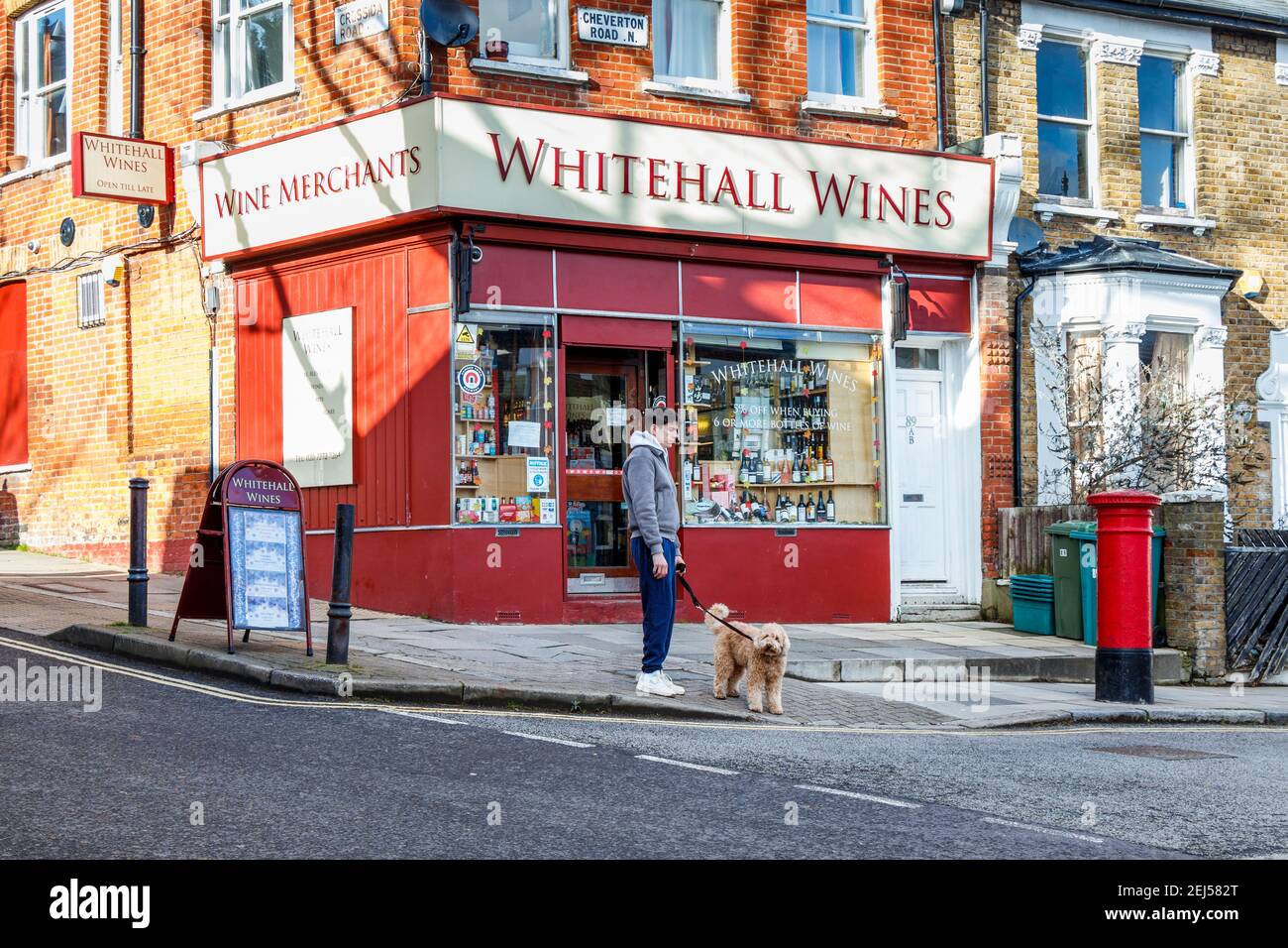 Ein Mann mit einem Hund an der Leine, der darauf wartet, die Straße außerhalb von Whitehall Wines zu überqueren, einem lokalen Lizenznehmer in Nord-London, Großbritannien Stockfoto