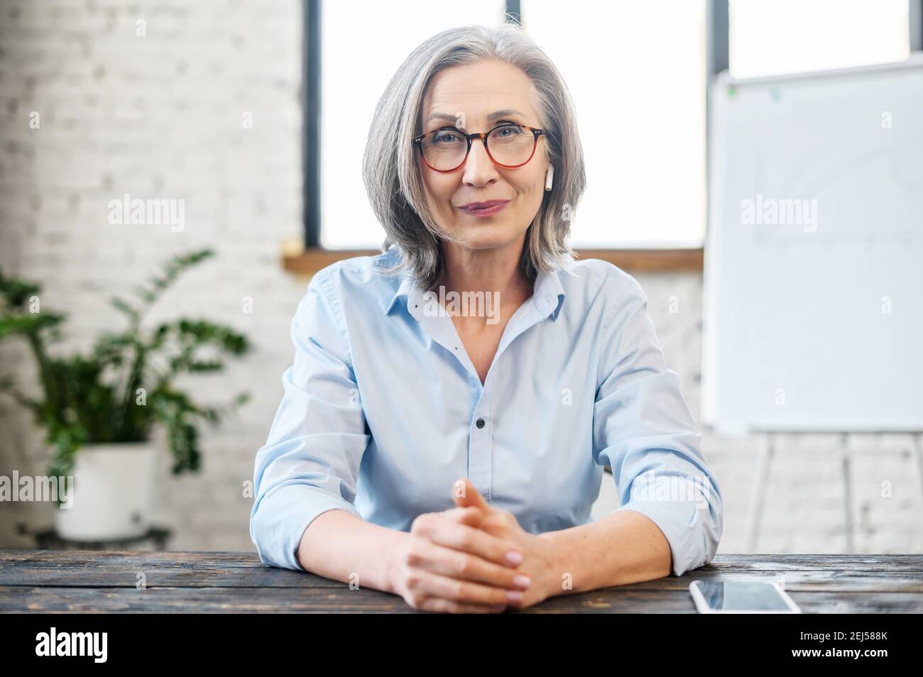 Serious intelligente ältere Lehrer Geschäftsfrau mit angenehmen Lächeln und Ohrhörer in Brille Blick auf Kamera, sitzen am Schreibtisch, bieten Stockfoto