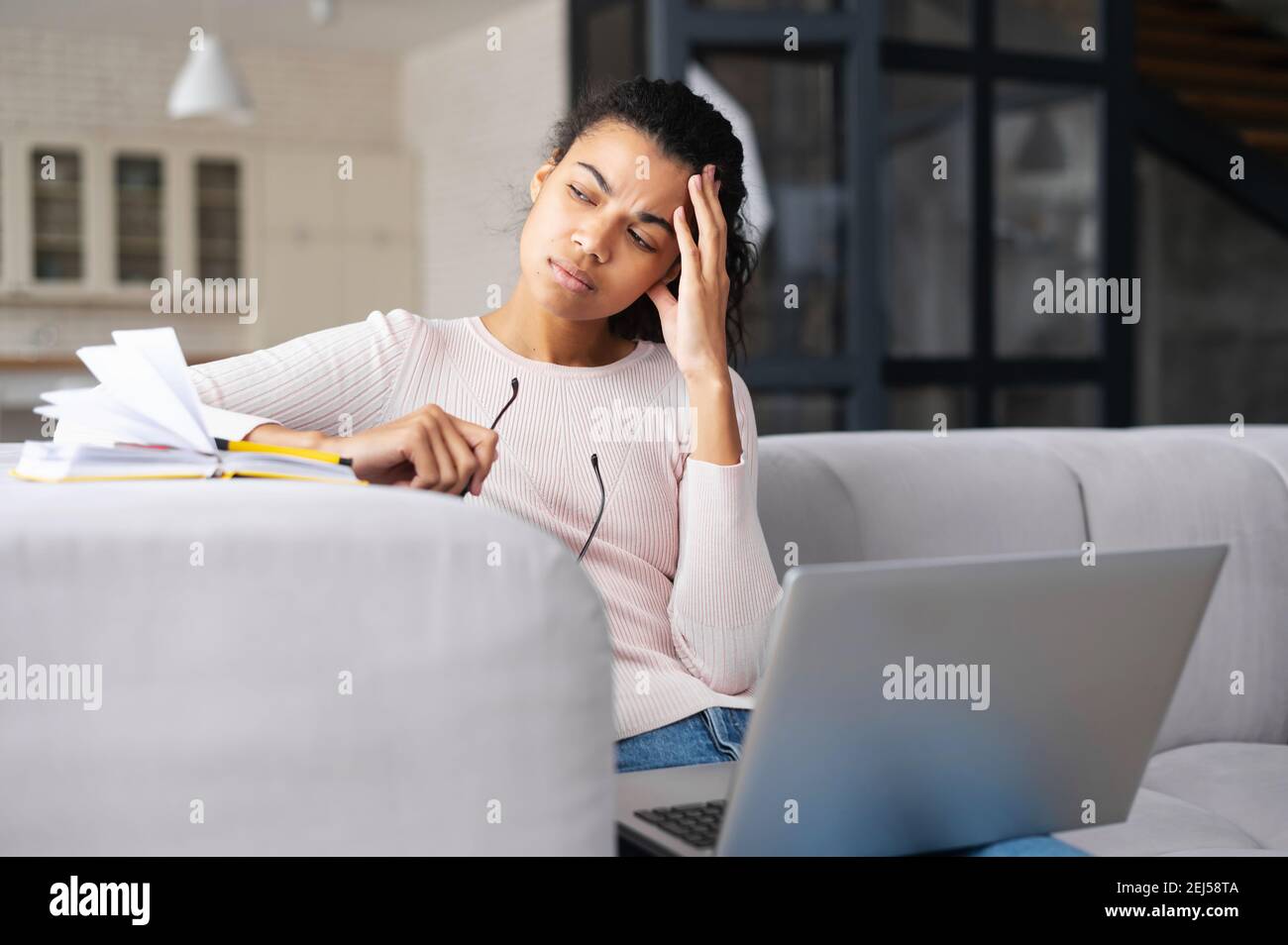 Müde multirassische junge Frau, die auf dem Sofa mit Laptop sitzt und wegschaut, faltige Stirn, Hand mit dem Kopf haltend, überarbeitet, hält Stockfoto