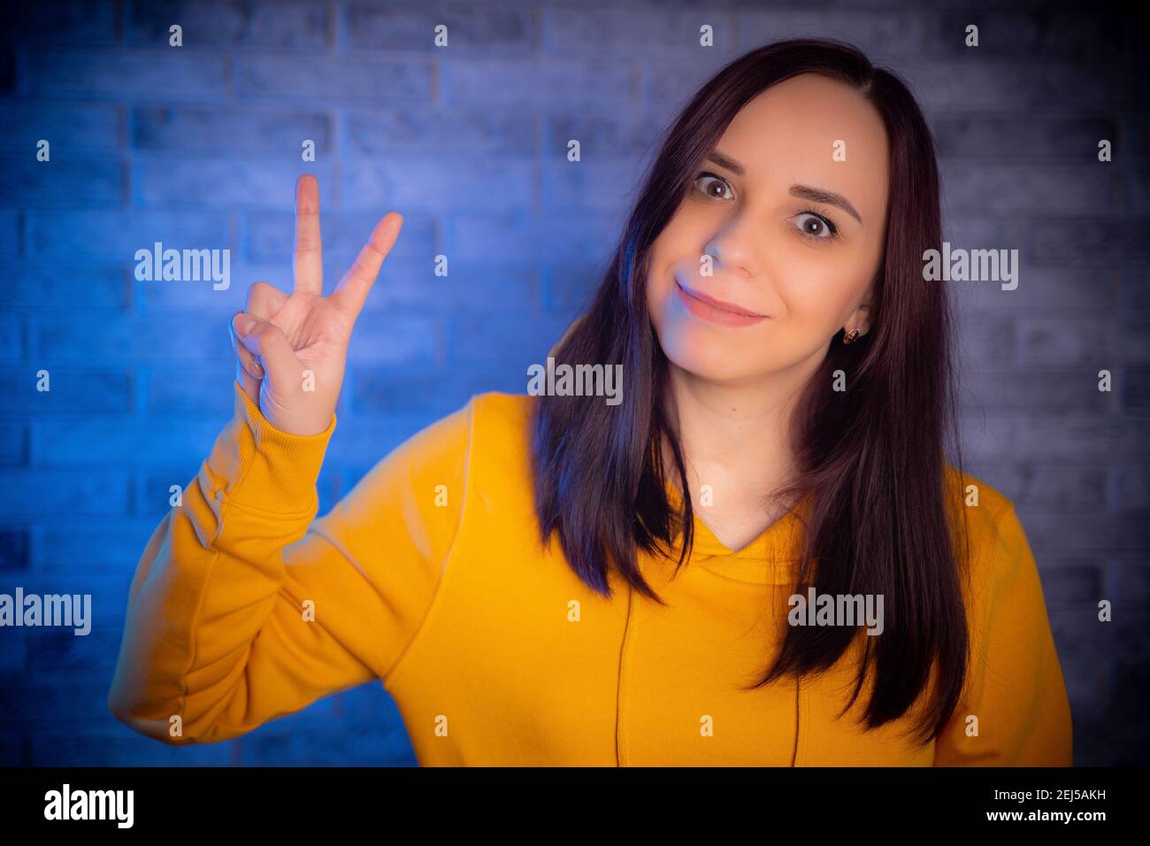 Porträt einer jungen Frau lächelnd und zeigt Daumen nach oben. Nahaufnahme der glücklichen Brünette zeigt Geste der Zustimmung auf hintergrundbeleuchteten Hintergrund Stockfoto