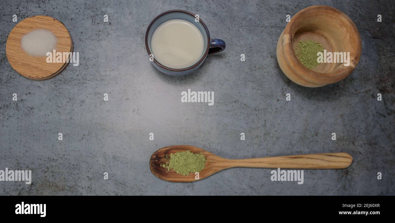 Draufsicht auf gesundes Matcha-Teepulver auf Holzlöffel mit einer Tasse Milch und Zucker in einem Grauen Stein Hintergrund. Matcha Tee Zubereitung Zutat 2021. Stockfoto