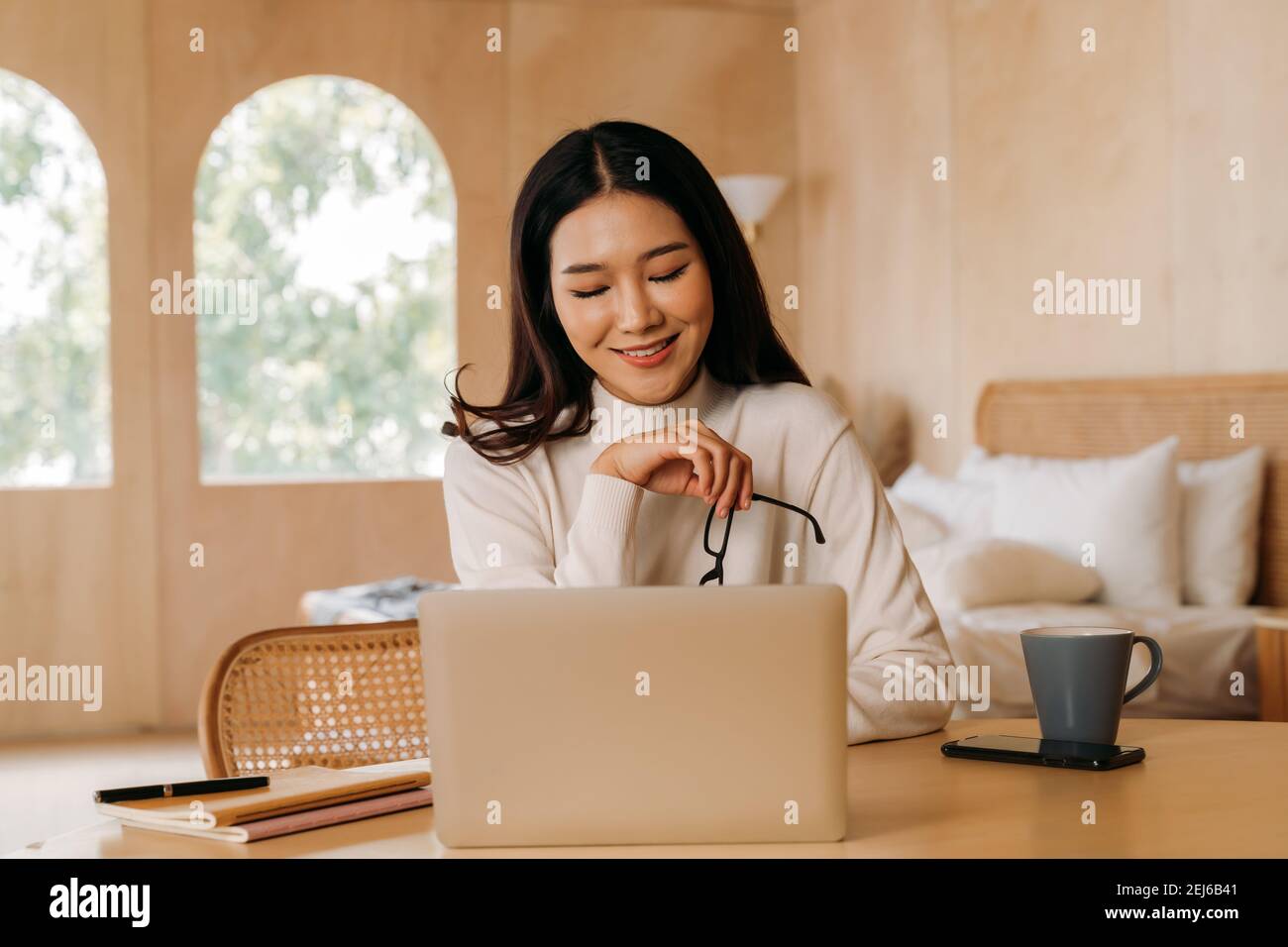 Junge asiatische Geschäftsfrau mit Brille und Laptop lächelnd beim Blick auf die Kamera. Freiberufliche Mitarbeiterin am Tisch sitzen und von zu Hause aus arbeiten. Stockfoto