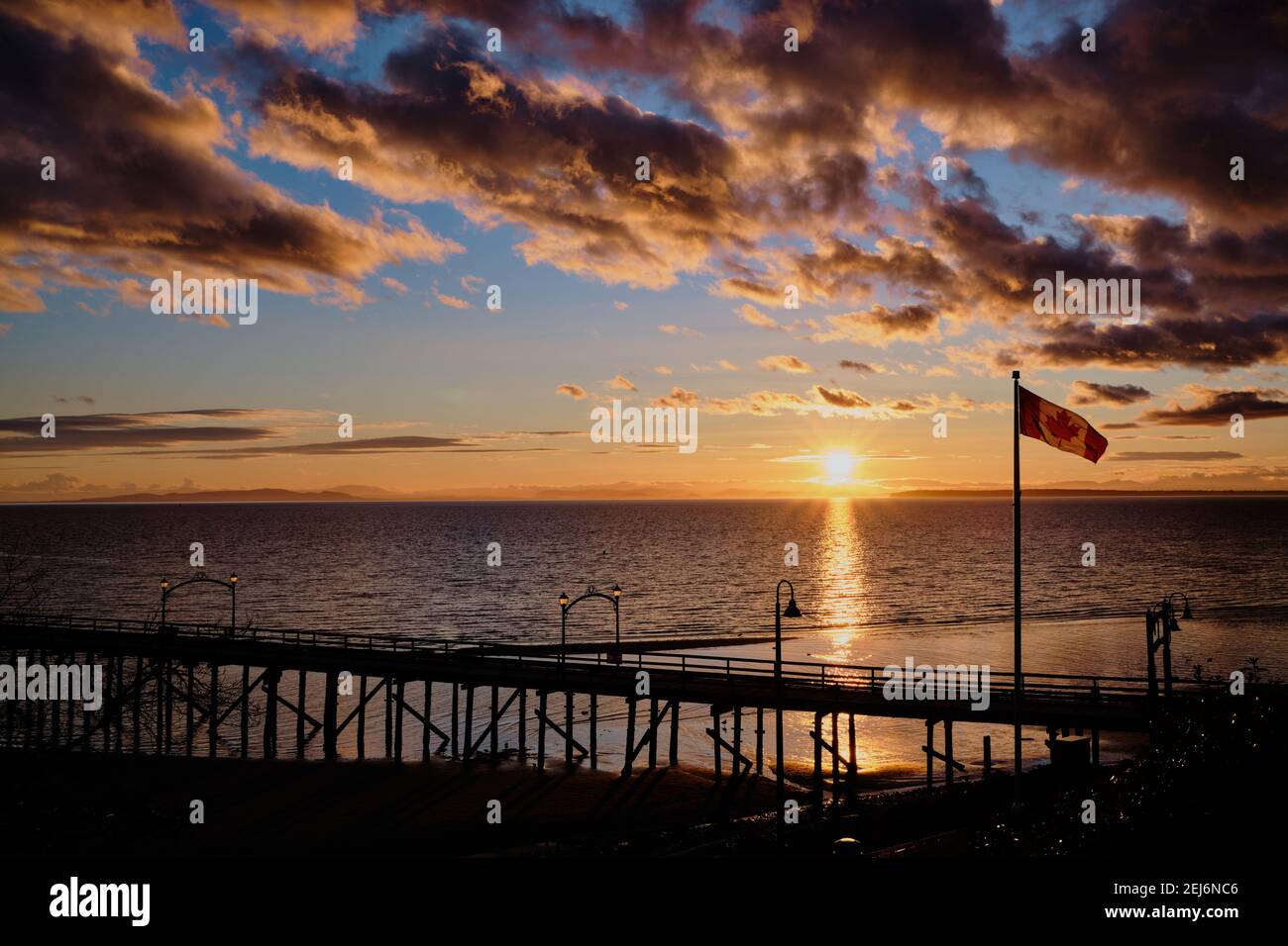 Spaziergänger auf dem berühmten Pier von White Rock genießen den atemberaubenden Sonnenuntergang über dem Meer. Im Vordergrund flattert Kanadas Nationalflagge stolz in der Brise. Stockfoto