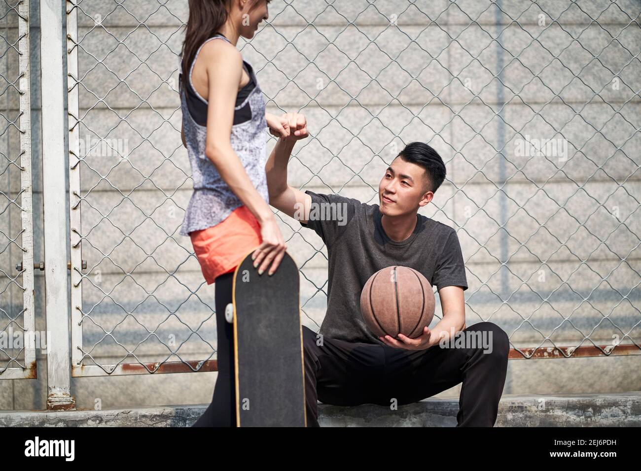 Junge asiatische Erwachsene männliche Basketballspieler und weibliche Skateboarder stoßen Fäuste auf dem Platz im Freien Stockfoto