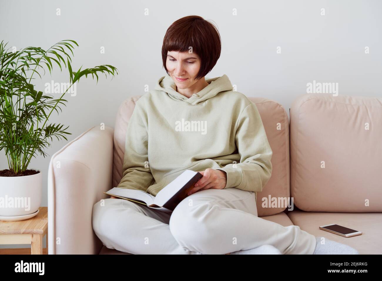 Reife Frau liest Buch sitzend auf Sofa, allein denkend. Digitales Detox-Konzept, Freizeit für sich Stockfoto
