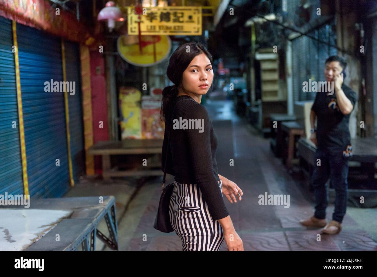 Junge schöne asiatische Mädchen in einer engen Gasse in der Nacht, zu Fuß weg von der Kamera, während der Blick zurück auf die Kamera. Mann, der im Hintergrund steht. Stockfoto