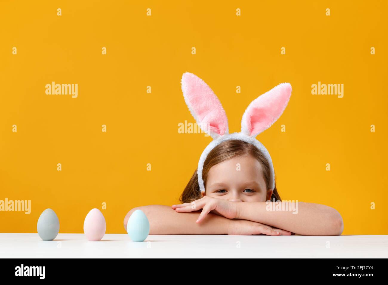 Nettes Kind in Osterhasen Ohren auf einem gelben Hintergrund. Ein kleines Mädchen schaut vom Tisch und jagt nach bemalten Eiern. Stockfoto