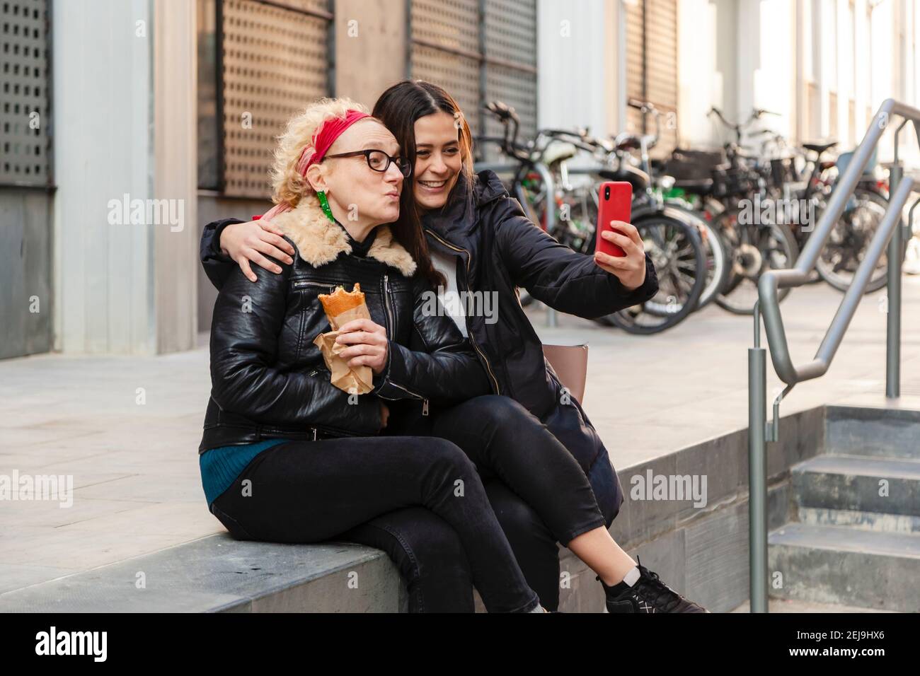 Zwei Freundinnen, die ein Selfie mit einem Smartphone festhalten, lächeln und in urbaner Umgebung eng beieinander sitzen Stockfoto