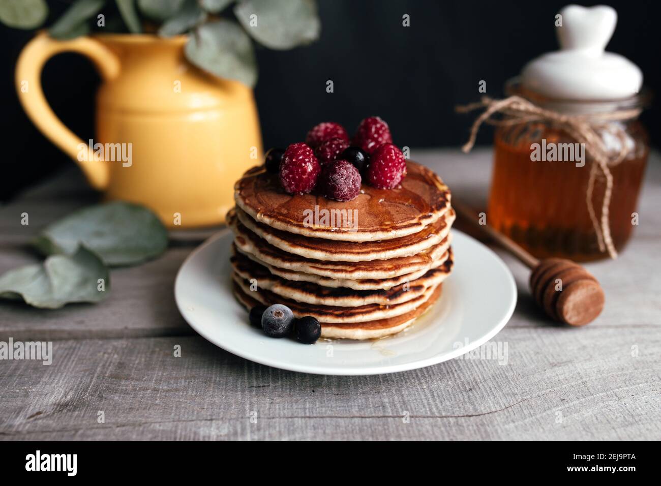 Saftige Pfannkuchen mit Beeren und Honig auf einem weißen Teller, Löffel, Glas, Holztisch, gelbe Vase mit Eukalyptus. Hochwertige Fotos Stockfoto