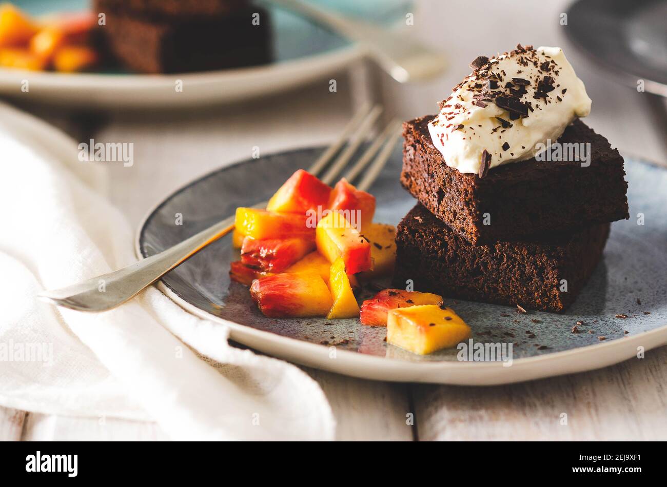 Brownies mit Sahne und Schokolade auf der Oberseite, Pfirsichstücke in hellblauen Tellern mit Gabeln und einer weißen Serviette, auf weißem Hintergrund. Stockfoto