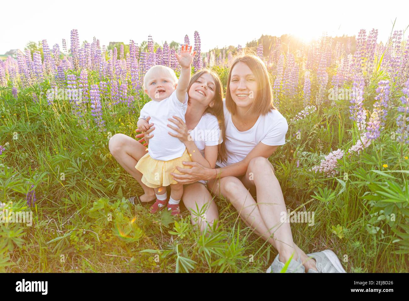 Junge Mutter umarmt ihre Kinder im Freien. Frau Baby Kind und Teenager-Mädchen sitzen auf Sommerfeld mit blühenden wilden Blumen grünen Hintergrund. Glücklich Stockfoto