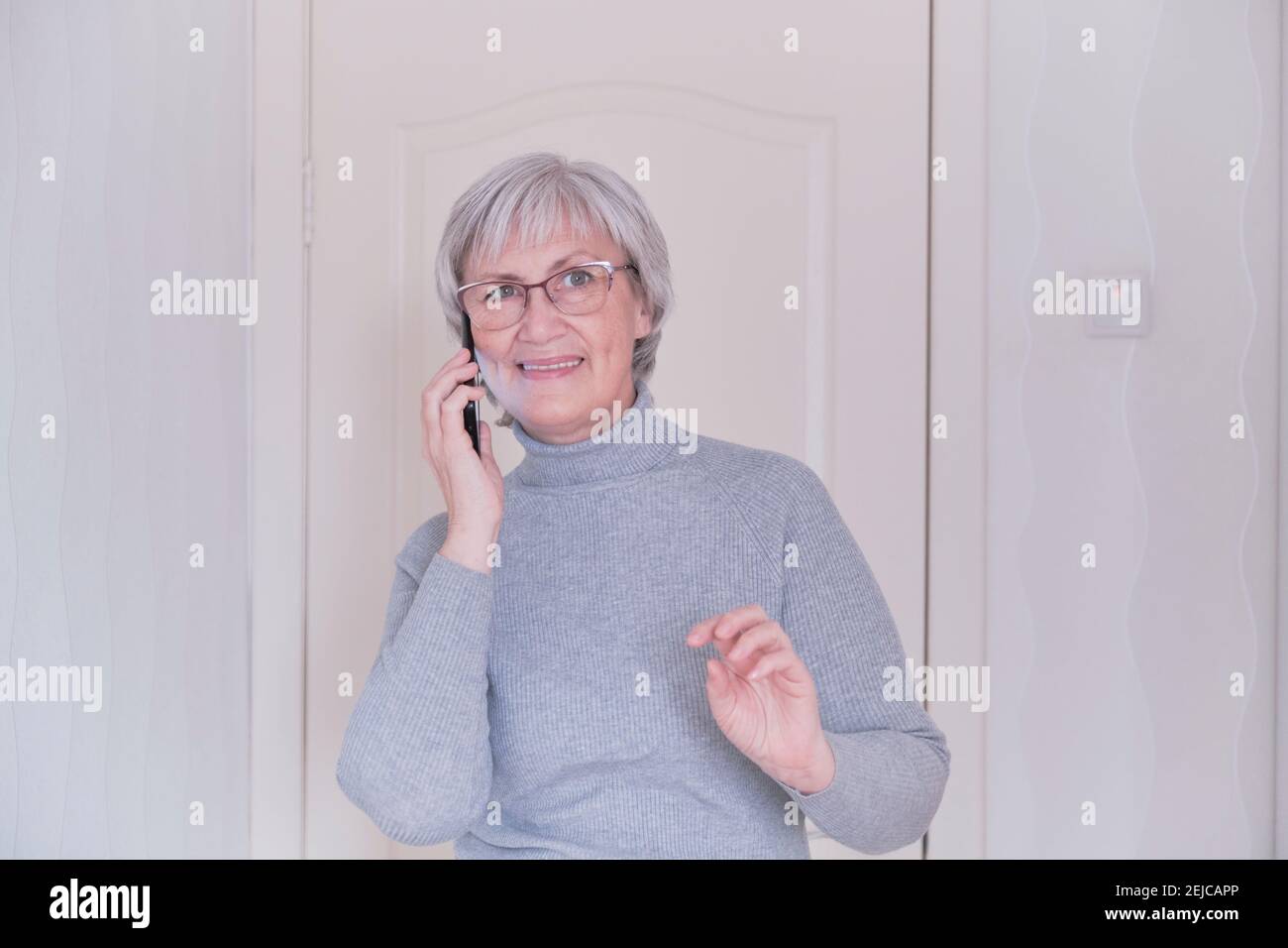 Eine grauhaarige ältere Frau mit Brille und einem grauen Rollkragen, die zu Hause auf dem Handy spricht. Menschen verbinden, Kommunikation. Lifestyle-Moment. Stockfoto