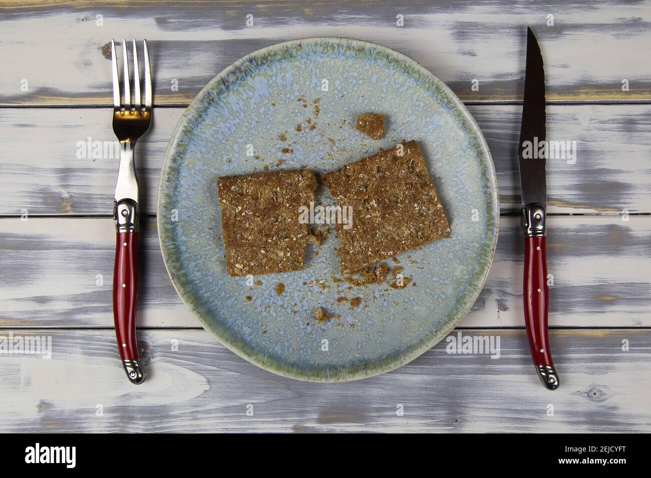 Draufsicht auf Teller mit einer Scheibe trocken zerbrochene Knäckebrot Krümel, Besteck auf Holztisch - unausgewogenes spärliches Diät-Konzept (Fokus auf Mitte) Stockfoto