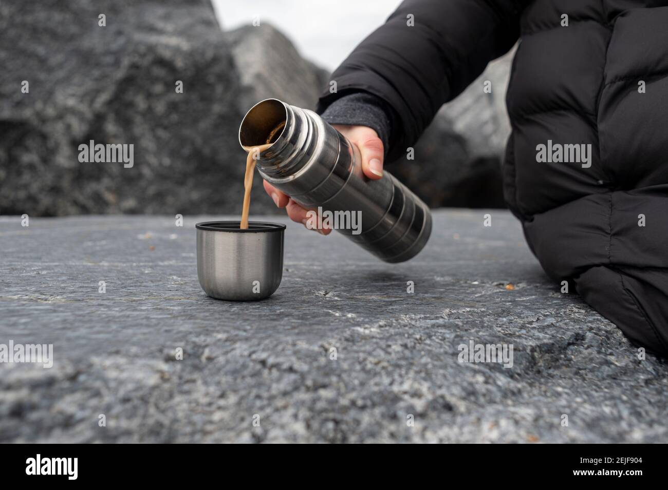 Zugeschnittenes Bild einer Frau, die auf Felsen sitzt und Thermoskannen hält, ein warmes Getränk in eine Tasse gießt und eine Pause macht, während sie in der Natur wandert. Stockfoto