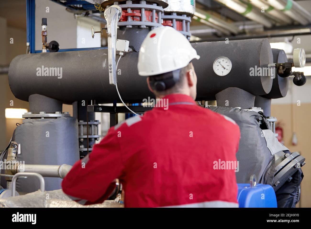 Thema der Energiewirtschaft: Ein Techniker in roten Overalls und einem weißen Helm, der die Heizungsparameter überprüft. Heizwasserverteilungstechnik. Stockfoto