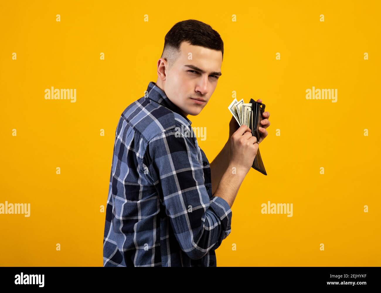 Junger Mann mit Geldbörse voller Geld, gierig über Sein Reichtum auf orangenen Studiohintergrund Stockfoto
