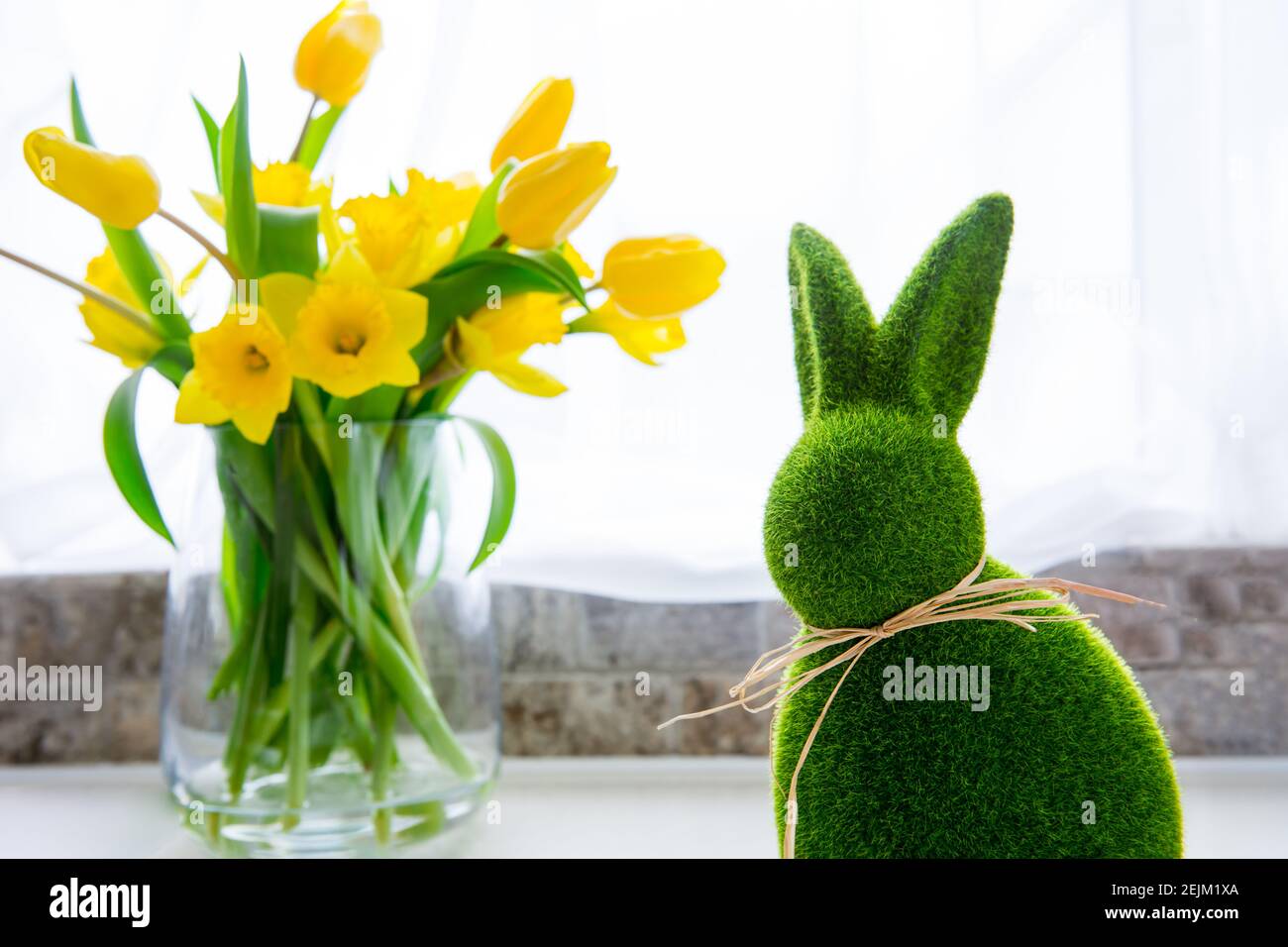 Niedliche grüne Gras Hase Kaninchen und Bouquet von frischen Frühling gelben Tulpen und Narzissen Blumen auf dem weißen Küchentisch in der Nähe des Fensters. Frohe Ostern Stockfoto