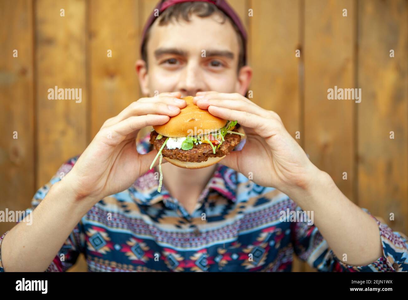 Kerl in einem stilvollen Shirt Kerl in einem stilvollen Shirt Mit einer Basiskappe genießen Sie einen schönen vegetarischen Burger auf Eine hölzerne Terrasse Stockfoto